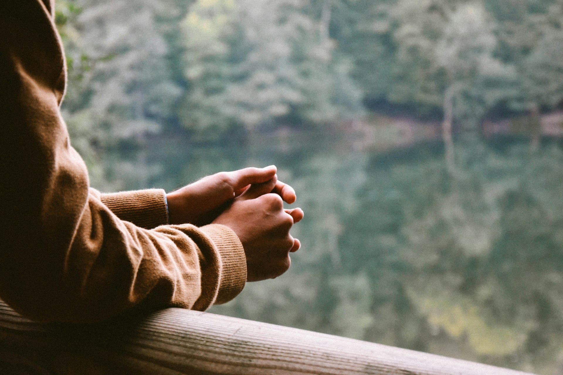 A person in a brown jumpers hands rested over wooden railing.
